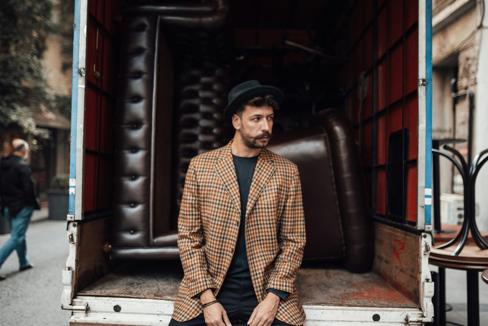 A fashionable man in a hat and checkered blazer leans against a truck with furniture inside.